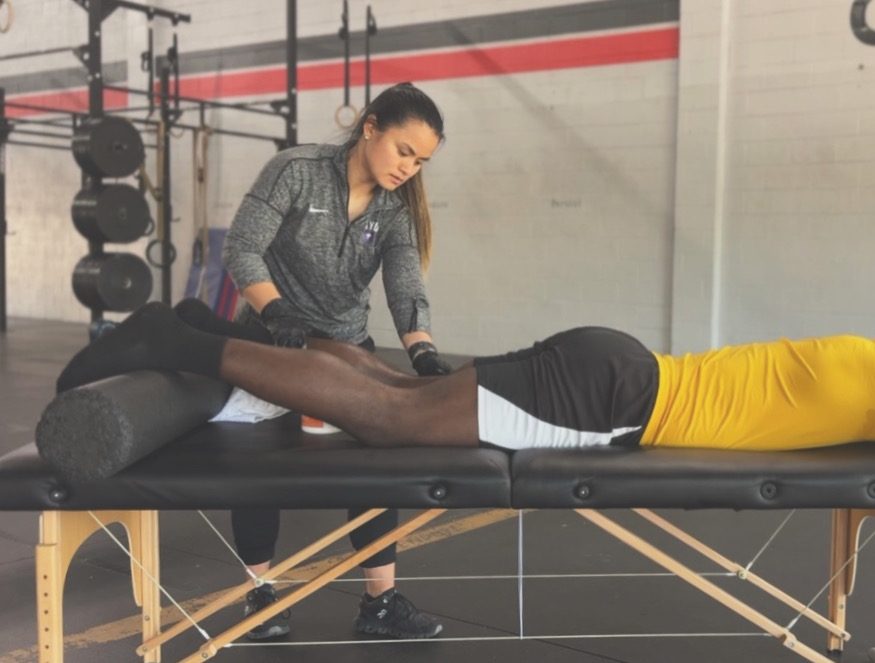Dr. Amy Driessen performing hands-on treatment on a patient's leg at a treatment table set up on the gym floor, with weight plates and a rig in the background.